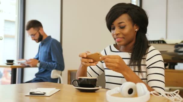 Retrato de una hermosa mujer africana bebiendo café y usando un teléfono inteligente mientras está sentada en la mesa en la cafetería moderna . — Vídeo de stock