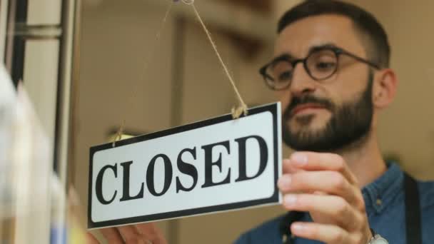 Egy fiatal férfi portréja a szemüvegben, amint egy "csukva" táblát fordít a kávézó ajtaján. Közelkép a pincérek kávézójának megnyitásáról.. — Stock videók