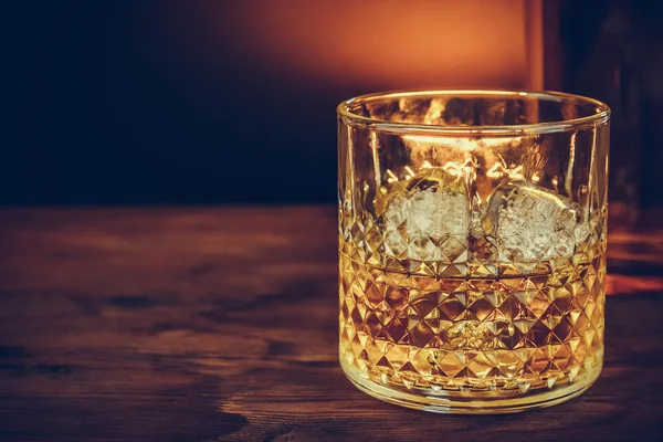Vaso de whisky con cubitos de hielo cerca de la botella en la mesa de madera, ambiente cálido — Foto de Stock