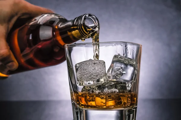 Barman pouring whiskey with ice cubes in glass on black background, cold atmosphere — Stock Photo, Image