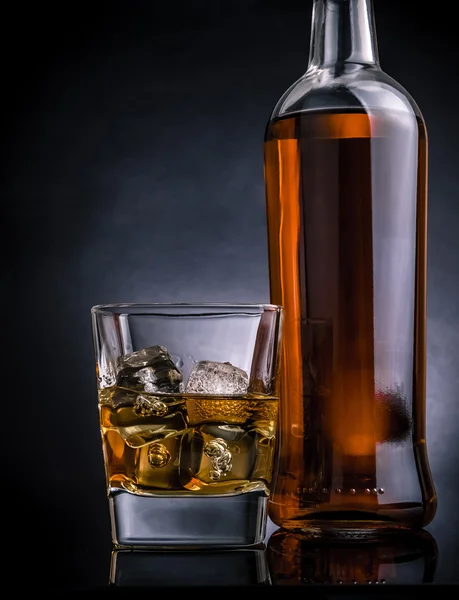Whisky s ledem ve skle poblíž láhev na černém pozadí, chladné atmosféře — Stock fotografie