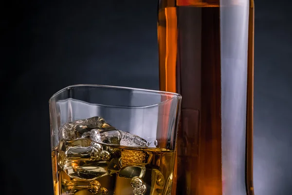 Närbild av whisky med isbitar i glaset nära flaskan på svart bakgrund, kall atmosfär — Stockfoto