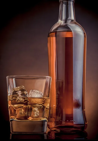 Whiskey with ice cubes in glass near bottle on black background, warm atmosphere — Stock Photo, Image