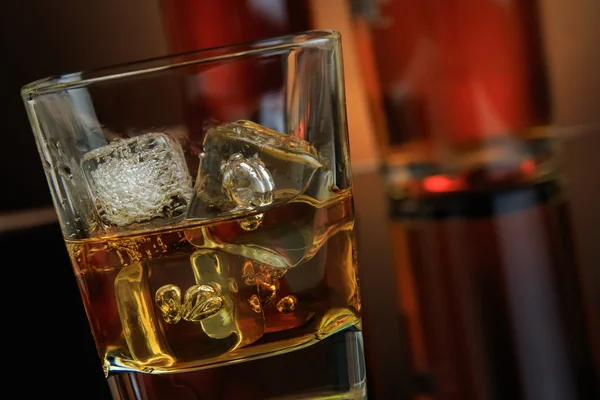 Close-up of whiskey with ice cubes in glass near bottle on black background, warm atmosphere — Stock Photo, Image