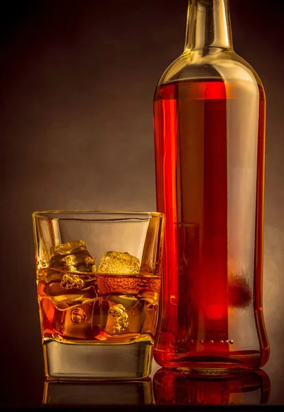 Whiskey with ice cubes in glass near bottle on black background, warm atmosphere — Stock Photo, Image
