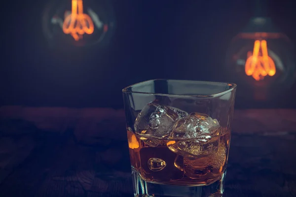 Vaso de whisky con cubitos de hielo en la mesa de madera, ambiente cálido, tiempo de relax con whisky con un poco de luz bombilla caliente — Foto de Stock