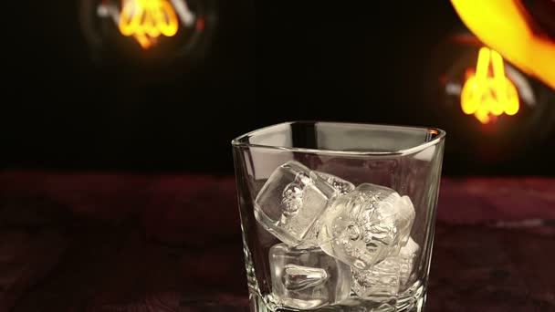 Barman versant du whisky avec des glaçons sur une table en bois, ambiance chaleureuse, moment de détente avec du whisky avec une ampoule vintage chaude et légère — Video