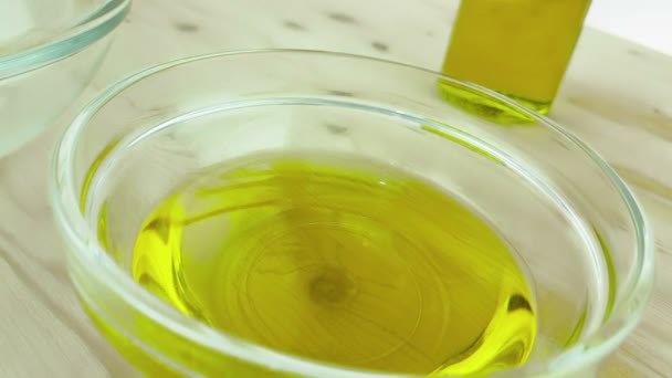 Verter el aceite de oliva de cocina en la botella en el recipiente blanco con gotas en el fondo de la mesa de madera cerca de la botella de aceite de oliva, disparado en cámara lenta en blanco, concepto de dieta de alimentos saludables — Vídeos de Stock