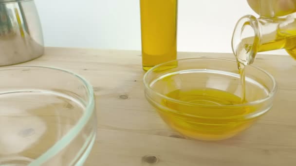 Versando l'olio di oliva in bottiglia nel contenitore bianco su sfondo tavolo di legno vicino alla bottiglia di olio d'oliva, girato al rallentatore su sfondo bianco, concetto di dieta alimentare sana — Video Stock