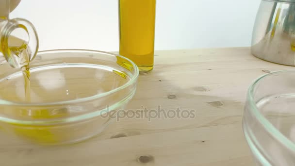 Versando l'olio di oliva in bottiglia nel contenitore bianco su sfondo tavolo di legno vicino alla bottiglia di olio d'oliva, girato al rallentatore su sfondo bianco, concetto di dieta alimentare sana — Video Stock