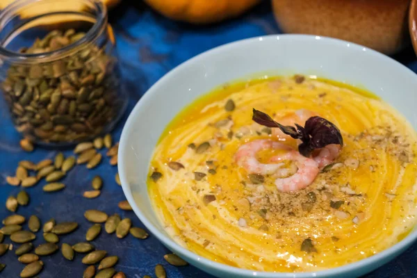 Autumn food - pumpkin soup with cream, shrimps and seeds in bowls on table. pumpkin soup decorated with pumpkin seeds and toasted crackers on a dark blue background. Served for dinner View from above — Stock Image Autumn Food Pumpkin Soup Cream Shrimps Seeds Bowls Table Pumpkin Stock Photo