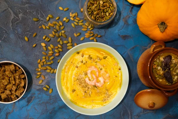 Autumn food - pumpkin soup with cream, shrimps and seeds in bowls on table. pumpkin soup decorated with pumpkin seeds and toasted crackers on a dark blue background. Served for dinner View from above — Stock Image Autumn Food Pumpkin Soup Cream Shrimps Seeds Bowls Table Pumpkin Stock Image
