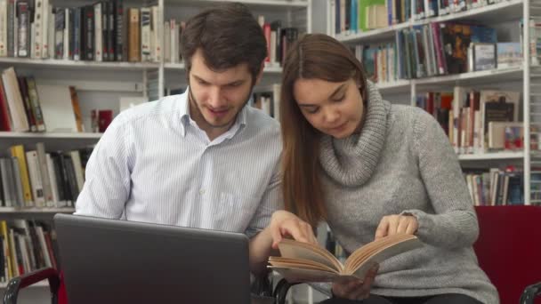 Uczniowie porównać informacje w książce i na laptopie w bibliotece — Wideo stockowe