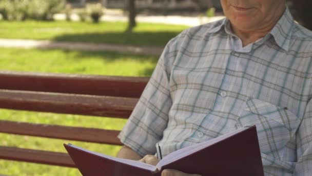 Hombre mayor escribe en su cuaderno en el banco — Vídeos de Stock