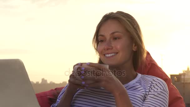 Uma menina descansando bebe chá quente e olha para o laptop sentado no telhado — Vídeo de Stock