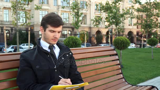 Ένα επιχειρησιακό άτομο που κάθεται σε ένα παγκάκι και γράφει κάτι — Αρχείο Βίντεο