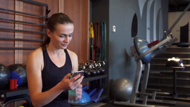 Спортивная девушка смотрит в телефон и пьет воду во время перерыва между упражнениями — стоковое видео