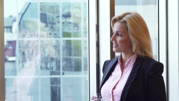 Vacker kvinna ser ut genom fönstret på sitt kontor och drycker vatten — Stockvideo