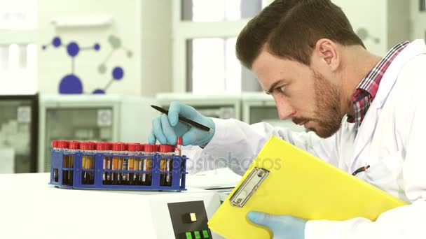 Un joven asistente de laboratorio saca tubos de ensayo con análisis y reescribe datos sobre ellos — Vídeos de Stock