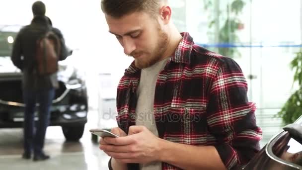 Hombre barbudo joven usando su teléfono inteligente en el concesionario de automóviles — Vídeo de stock