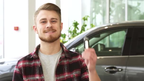 Atractivo joven sonriendo sosteniendo las llaves del coche posando en el concesionario de automóviles — Vídeos de Stock