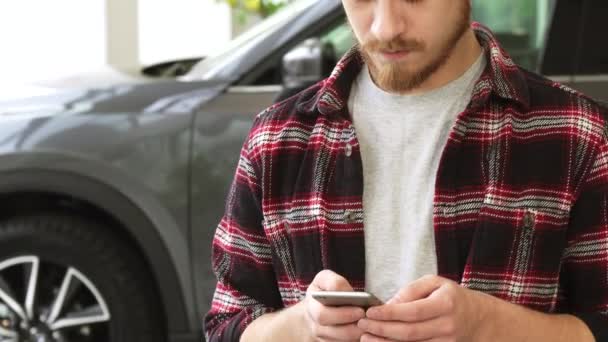 Skäggiga stilig ung man med hjälp av smartphone samtidigt köpa ny bil på generalagenten — Stockvideo