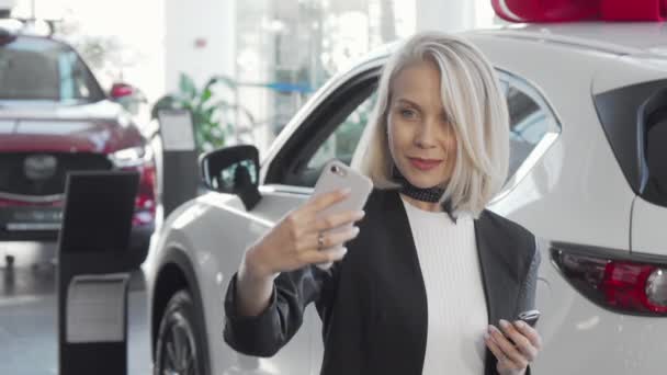 Mulher feliz tirando selfies com chaves para seu carro novo na concessionária — Vídeo de Stock