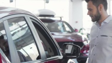 Satılık arabayı incelerken kameraya gülümseyen yakışıklı bir adam.