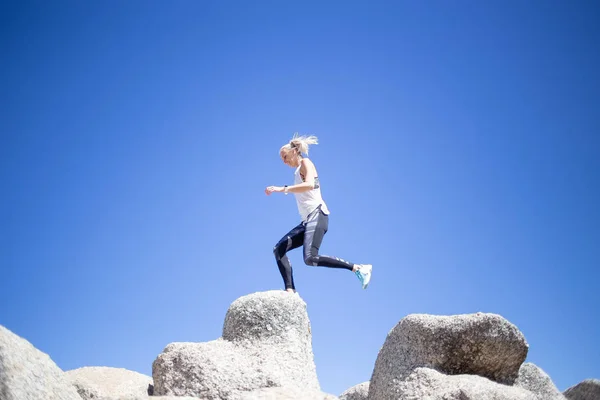 Beautiful blond model in sportswear jumping from rock to rock against bright blue sky at daytime