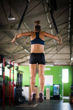 Spor salonunda kadın fitness modeli kendini yukarı çekerek kafa halkaları üzerinde çapraz antrenman yapıyor.