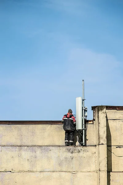 Un travailleur qui fixe une antenne radio GSM LTE cellulaire sur le dessus du bâtiment. Fond bleu ciel — Photo