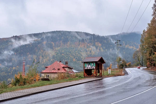 Fog and mist over the pine mountain forest with traditional old village houses, a red car on the road and a fancy bus stop. — Stock Photo, Image