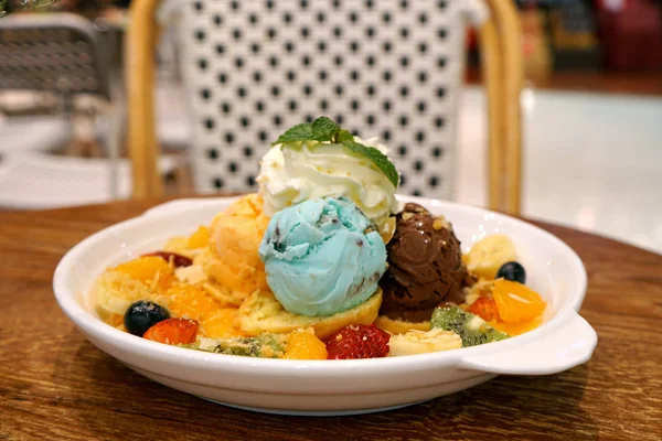 Assorted flavors scoop ice cream with giwi, strawberry, orange, blueberry, banana fruit and topping dress whip cream and mint leaves in white plate on wooden table at restaurant