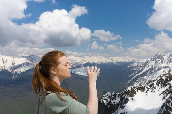 Young woman drink water from plastic bottle in the mountains on the snow peaks bacground