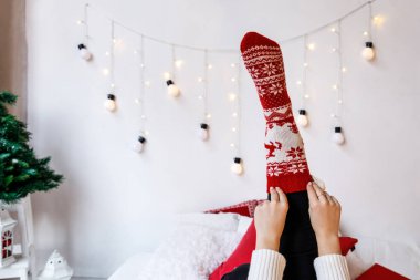 Beyaz duvarlı odada, kırmızı Noel süslemeleri ve ışıkları olan sıcak örülü çoraplar giyen hoş bir kadın. Tatil, yatak konseptinde rahatla.