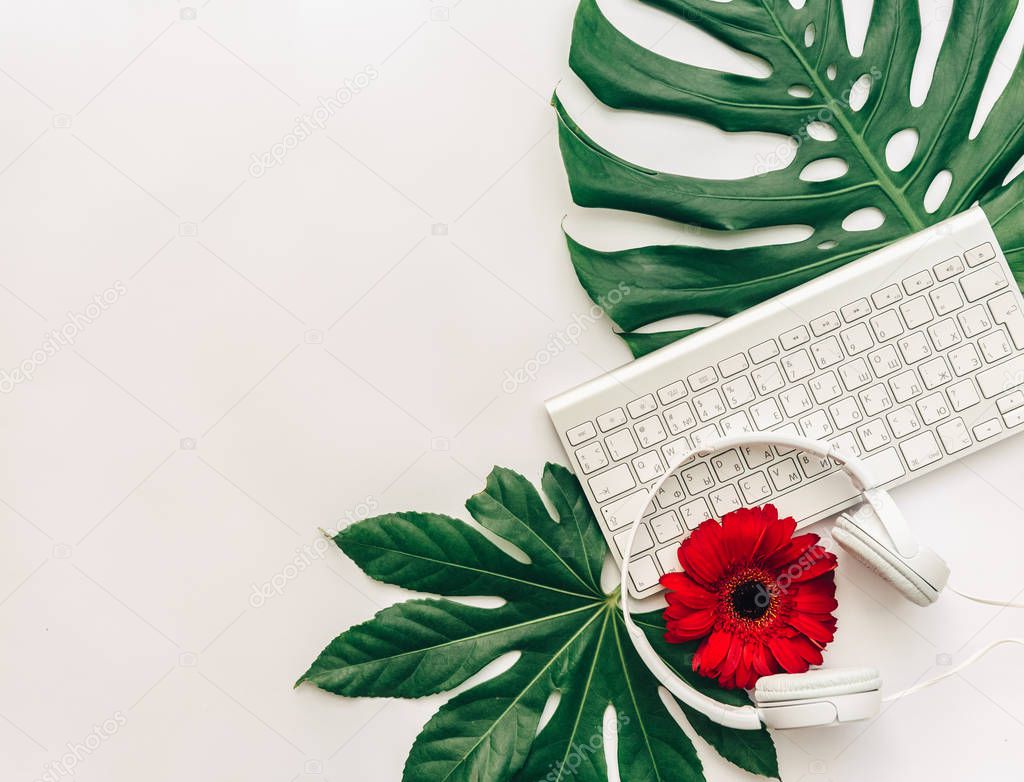 Creative flat lay with keyboard, headphones over tropical leaf monstera with flowers