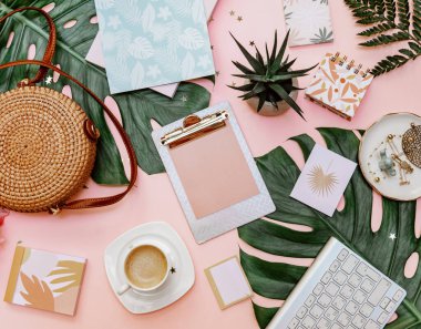 Kadın ofis masası dümdüz uzanıyordu. Klipsli kadın çalışma alanı, tropikal yapraklar, aksesuarlar, pembe arka planda bir fincan kahve. Üst görünüm. Boşluğu kopyala