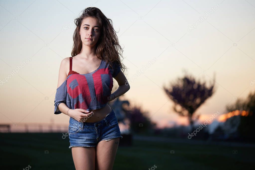 Beautiful brunette young woman with shortsand top posing above sunset background