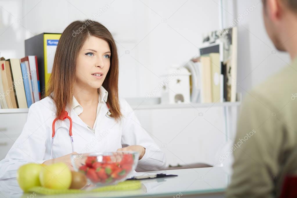 Smiling doctor nutritionist giving kiwi to male patient sitting at the desk with colorful fruits and vegetables