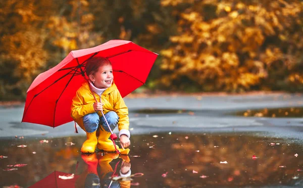 Happy child girl with umbrella and paper boat in   puddle in   a — Stock Photo, Image