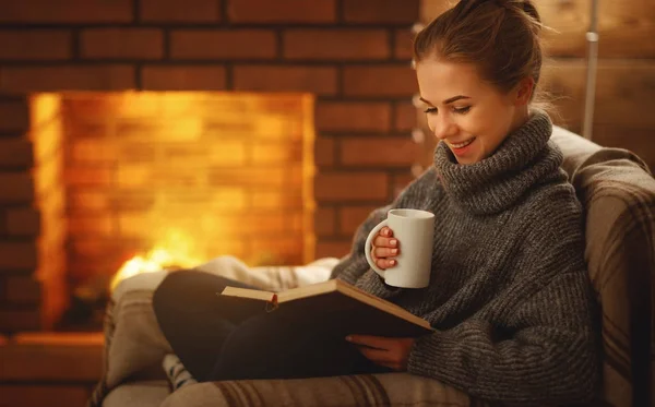 Ung kvinna läsa en bok vid den öppna spisen på en vinter evenin — Stockfoto