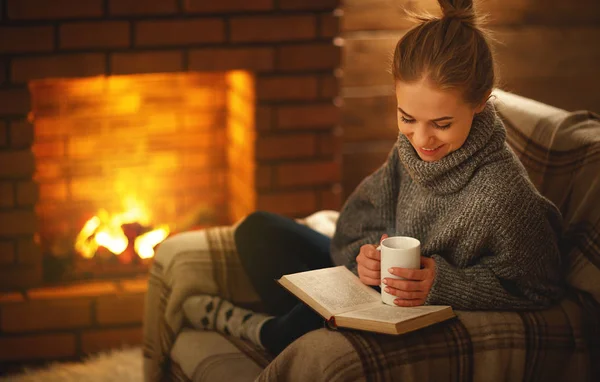 Jovem mulher lendo um livro junto à lareira em um anoitecer de inverno — Fotografia de Stock