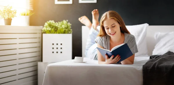 Happy young woman reads  book and drinks coffee in bed — Stock Photo, Image