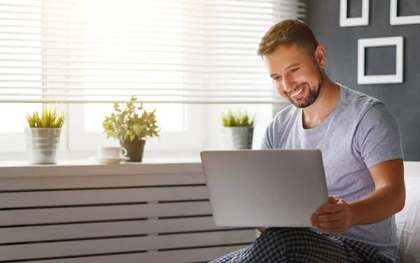 Glücklicher junger Mann mit Laptop und Tasse Kaffee im Bett — Stockfoto