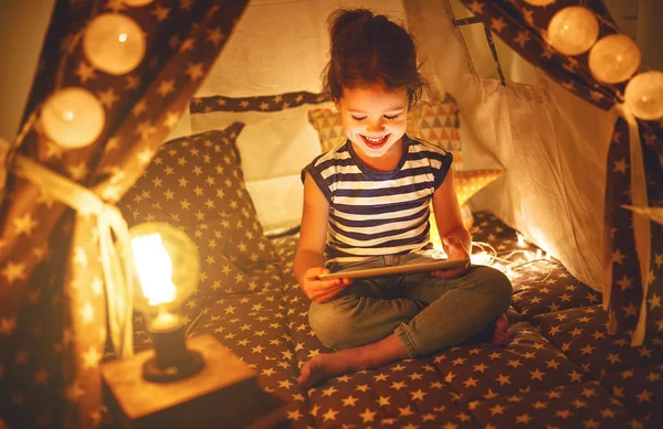 Gadis kecil yang bahagia dengan tablet pc tertawa dalam gelap di tenda di rumah — Stok Foto