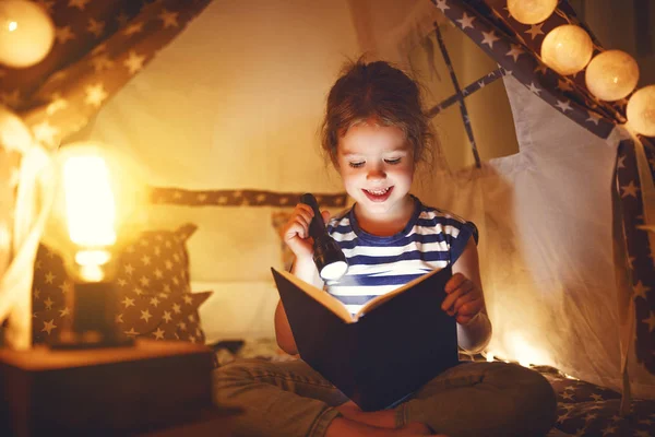 Feliz niña riendo y leyendo libro en la oscuridad en la tienda de campaña en ho — Foto de Stock