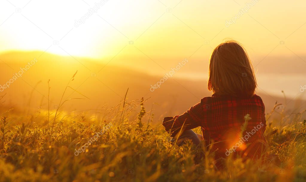 Woman sits with her back in the field and look sunset in the mou