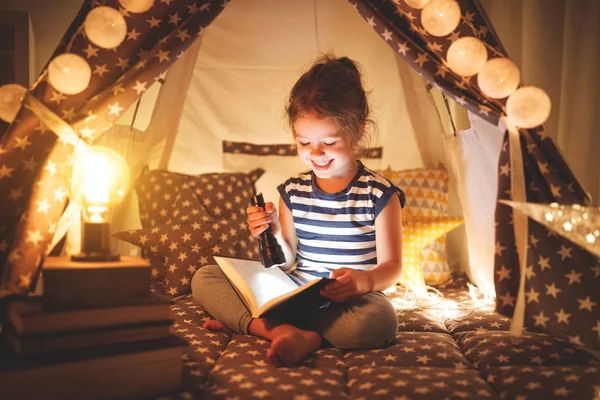 Feliz niña riendo y leyendo libro en la oscuridad en la tienda de campaña en ho — Foto de Stock