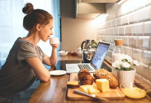 Kvinna i pyjamas har frukost vid datorn i kök — Stockfoto