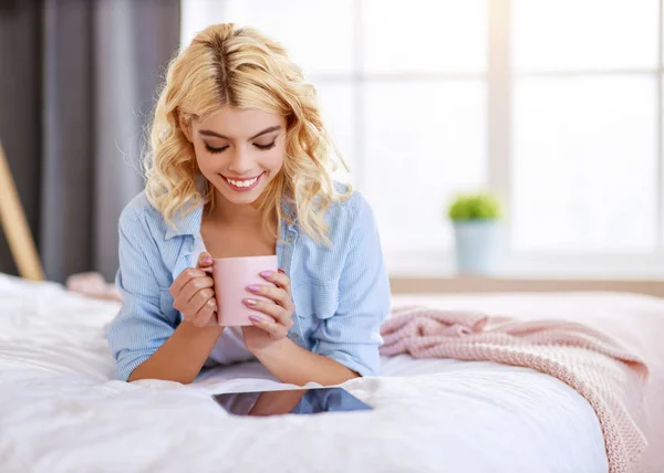 快乐的女孩一边喝咖啡，一边在床上用平板电脑笑 — 图库照片